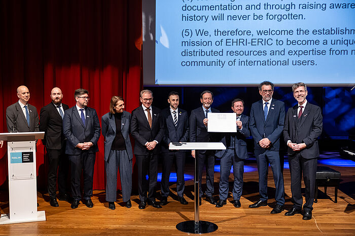 Gruppenfoto mit neun Männern und einer Frau auf einer Bühne vor rotem Vorhang und einer Leinwand, auf der englischsprachiger Text zu lesen ist.